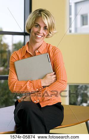Portrait of a businesswoman sitting on a desk holding a laptop View Large Photo Image Picture - Portrait of a businesswoman sitting on a desk holding a laptop. Fotosearch