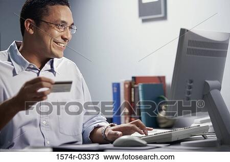 Mid adult man sitting in front of a computer holding a credit card View Large Photo Image Stock Image - Mid adult man sitting in front of a computer holding a credit card. Fotosearch