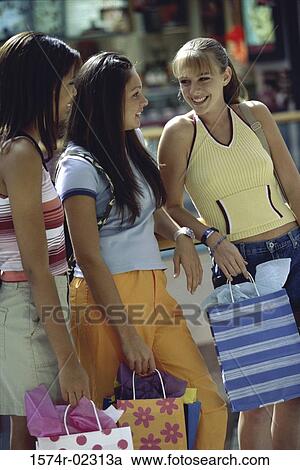 Stock Image - Side profile of three teenage girls standing with shopping bags. Fotosearch