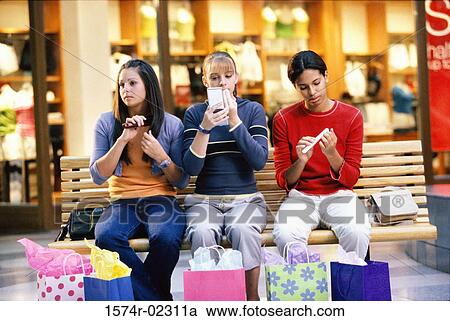 Stock Image - Three teenage girls sitting on a bench in a shopping mall applying make-up. Fotosearch