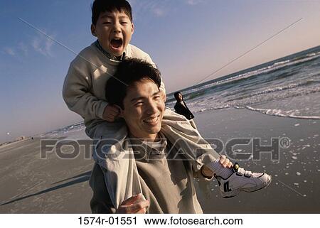 Stock Image - Close-up of a man carrying his son on his shoulders at the beach. Fotosearch