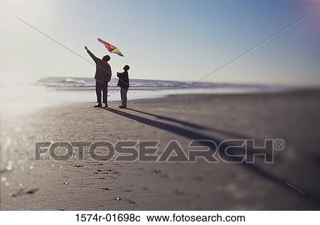 Stock Image - Silhouette of a father and son flying a kite on the beach. Fotosearch