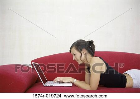 Side profile of a young woman lying on a couch and using a laptop View Large Photo Image Picture - Side profile of a young woman lying on a couch and using a laptop. Fotosearch