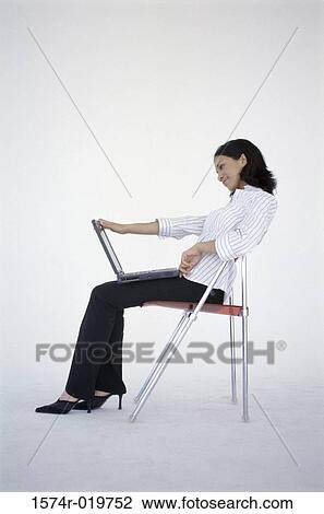 Stock Image - Side profile of a young woman sitting in a chair with a laptop on her lap. Fotosearch