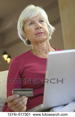 Stock Image - Low angle view of a senior woman sitting in front of a laptop holding a credit card. Fotosearch