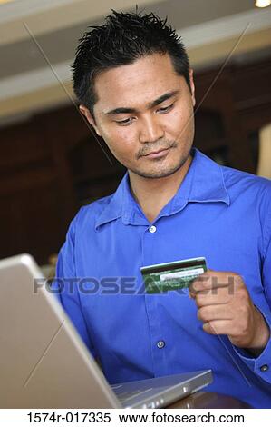 Low angle view of a young man sitting in front of a laptop looking at a credit card View Large Photo Image Stock Photography - Low angle view of a young man sitting in front of a laptop looking at a credit card. Fotosearch