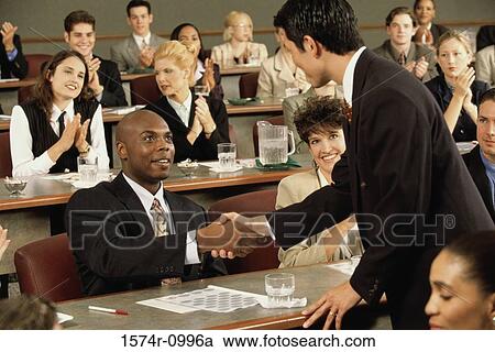 Stock Image - Businessmen shaking hands with people clapping in the background. Fotosearch