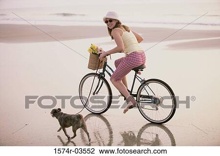Stock Image - Portrait of a young woman riding a bicycle with her dog running alongside. Fotosearch