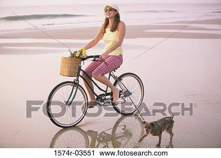 Stock Image - Young woman riding a bicycle with her dog running alongside. Fotosearch