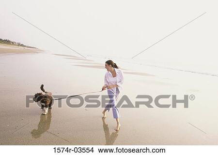 Picture - Young woman taking her dog for a walk on the beach. Fotosearch