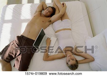 Stock Photo - High angle view of a young couple lying on the bed with a laptop. Fotosearch