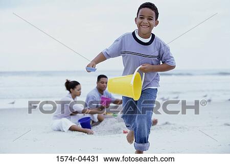 Stock Image - Boy playing on the beach with his parents sitting behind him. Fotosearch