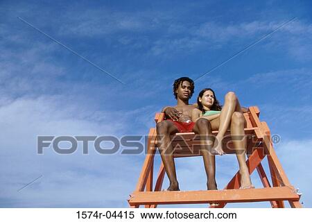 Stock Photography - Low angle view of a young couple sitting in a lifeguard chair on the beach. Fotosearch