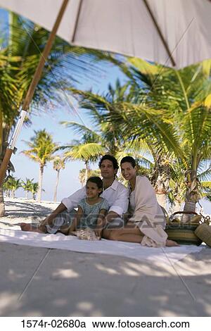 Stock Image - Low angle view of parents sitting with their daughter on the beach. Fotosearch