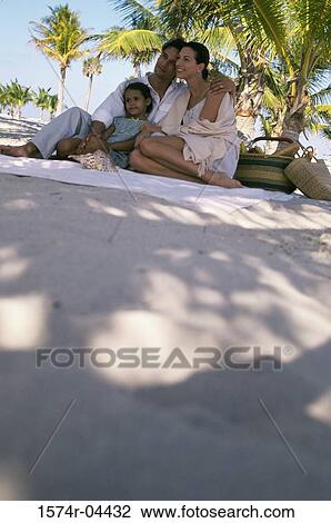 Stock Image - Low angle view of parents sitting with their daughter on the beach. Fotosearch