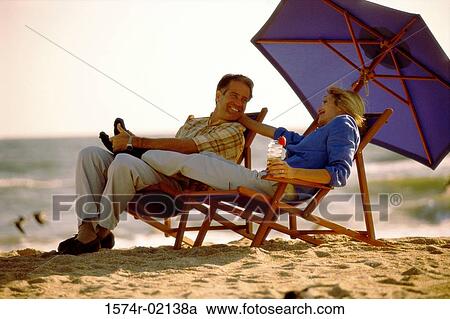 Mid adult couple sitting on lounge chairs on the beach View Large Photo Image Stock Image - Mid adult couple sitting on lounge chairs on the beach. Fotosearch