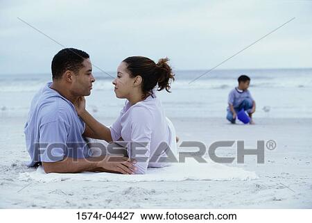 Stock Photo - Parents lying on the beach with their son playing behind them. Fotosearch