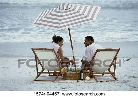 Stock Photo - Rear view of a young couple sitting in chairs on the beach. Fotosearch