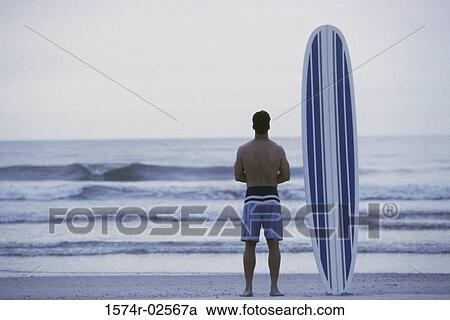 Rear view of a young man holding a surfboard standing on the beach View Large Photo Image Stock Image - Rear view of a young man holding a surfboard standing on the beach. Fotosearch