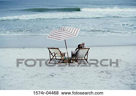Stock Photo - Rear view of a young man sitting on a deck chair on the beach. Fotosearch
