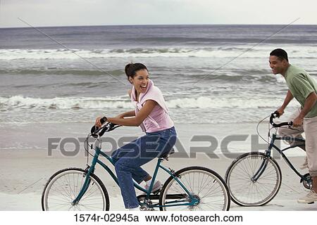 Stock Image - Side profile of a young couple cycling on the beach. Fotosearch