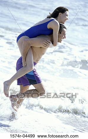 Stock Photo - Side profile of a young woman riding piggyback on a young man on the beach. Fotosearch
