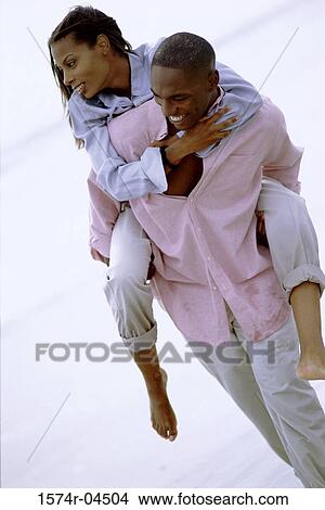 Picture - Young woman riding piggyback on a young man on the beach. Fotosearch
