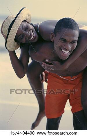 Stock Image - Young woman riding piggyback on a young man on the beach. Fotosearch