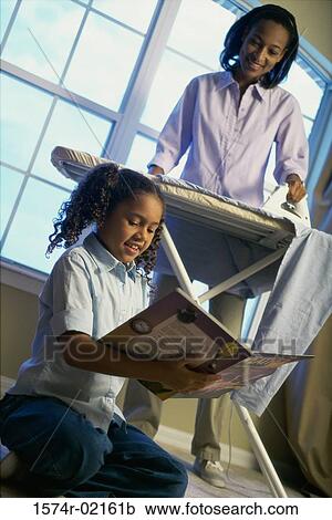 Stock Image - Mother ironing clothes with her daughter sitting on floor reading. Fotosearch