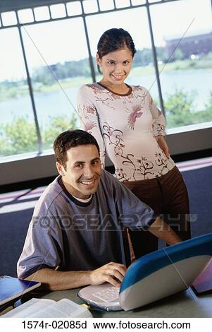 Stock Image - Portrait of a young man sitting in front of a laptop with a young woman standing behind him. Fotosearch