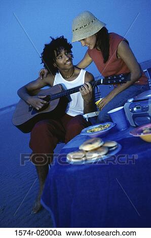 Young man playing a guitar on the beach with a young woman standing behind him View Large Photo Image Stock Image - Young man playing a guitar on the beach with a young woman standing behind him. Fotosearch