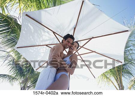 Stock Image - Portrait of a young couple standing under an umbrella smiling. Fotosearch