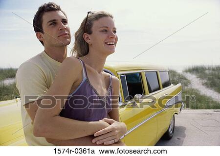 Stock Image - Young man leaning against a car holding a young woman from behind. Fotosearch
