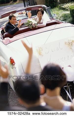 Stock Image - High angle view of a newlywed couple waving from a convertible car. Fotosearch