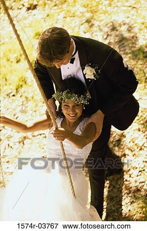 Stock Photo - Portrait of newlywed young woman sitting on a swing with a her groom pushing her. Fotosearch