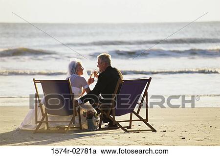 Stock Image - Senior couple holding glasses of champagne on the beach. Fotosearch