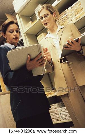 Stock Image - Low angle view of two businesswomen looking at files in a supply room. Fotosearch