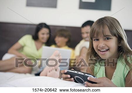 Portrait of a girl holding a handheld video game with her parents and brother looking at a laptop in the background View Large Photo Image Stock Image - Portrait of a girl holding a handheld video game with her parents and brother looking at a laptop in the background. Fotosearch
