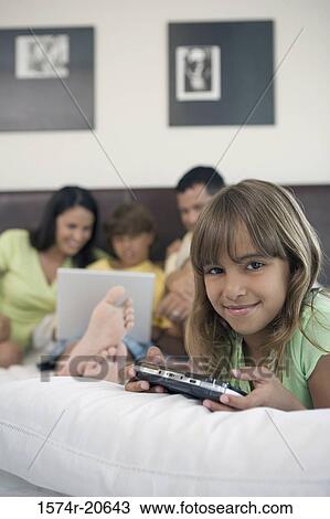 Stock Image - Portrait of a girl holding a handheld video game with her parents and brother looking at a laptop in the background. Fotosearch
