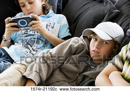 Stock Image - Portrait of a boy lying on the bed with another boy playing a handheld video game beside him. Fotosearch