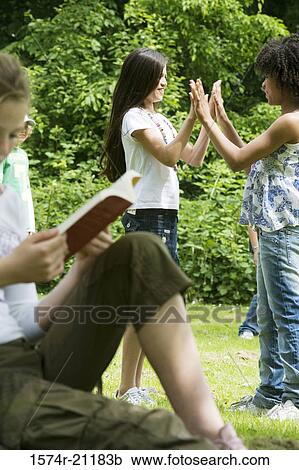 Stock Image - Side profile of a girl reading a book with two girls playing in the background. Fotosearch