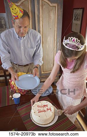 Stock Image - High angle view of a teenage girl cutting a birthday cake with her grandfather standing near her. Fotosearch
