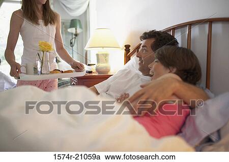 Stock Image - Side profile of a mid adult couple in the bed with their daughter holding a tray standing beside the bed. Fotosearch