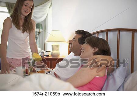 Stock Image - Side profile of a mid adult couple in the bed with their daughter holding a tray standing beside the bed. Fotosearch