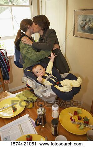 Stock Image - Side profile of a mid adult woman kissing her daughter with her son in a baby seat beside them. Fotosearch