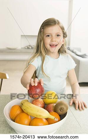 Stock Image - Portrait of a girl picking up an apple from a fruit bowl. Fotosearch