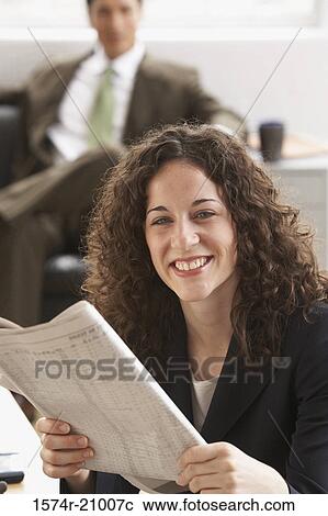 Stock Image - Close-up of a businesswoman holding a newspaper with a businessman sitting in the background. Fotosearch
