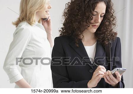 Close-up of a businesswoman using a PDA with another businesswoman talking on a mobile phone View Large Photo Image Stock Image - Close-up of a businesswoman using a PDA with another businesswoman talking on a mobile phone. Fotosearch