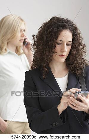 Stock Image - Close-up of a businesswoman using a PDA with another businesswoman talking on a mobile phone. Fotosearch