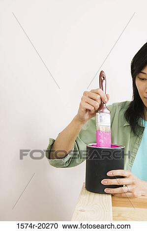 Stock Image - Close-up of a young woman dipping a paintbrush into a paint can. Fotosearch